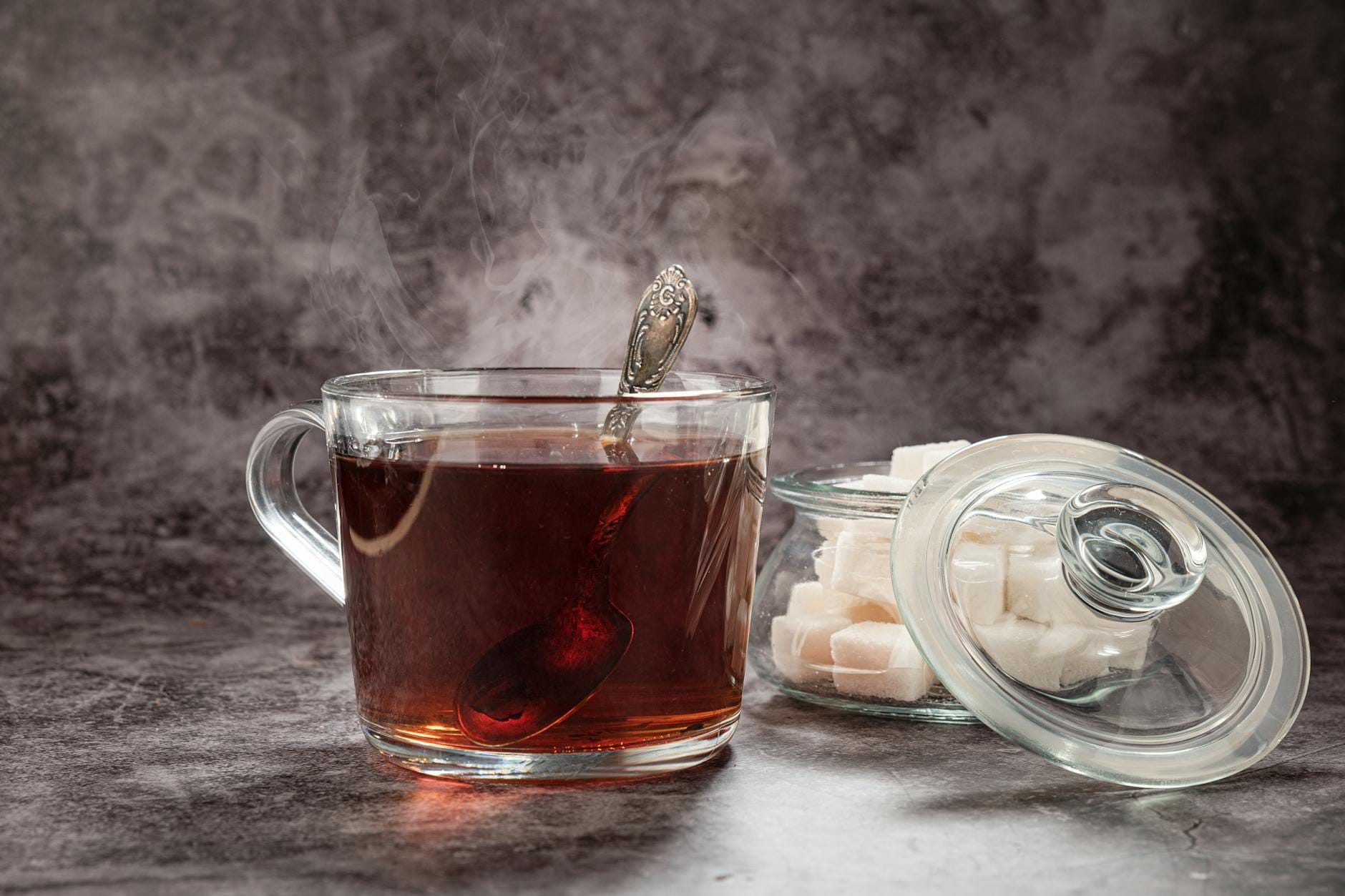 Gorąca herbata z parującą łyżeczką obok słoika z kostkami cukru